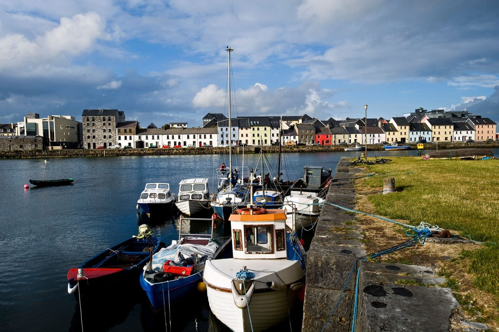 Eight fishing boats are tied up to a concrete quay, separating the sea from the land. In the background you can see houses, lined in rows next to each other of various colors of white, blue, red, and muted yellow and orange.